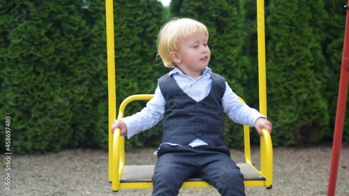 Happy light-haired toddler is swaying on swing in park at summer day, portrait of happy and laughing child. Adorable cute baby infant boy child at playground swinging.