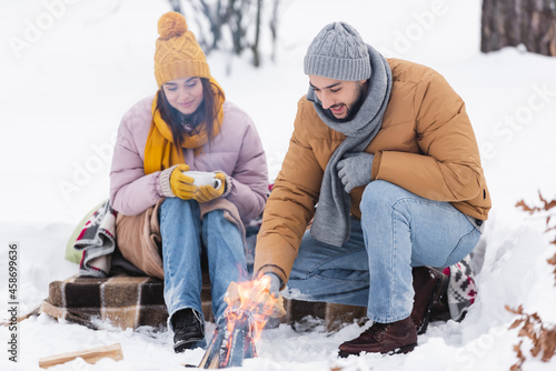Φωτογραφία Smiling man holding log near bonfire and girlfriend with cup in winter park