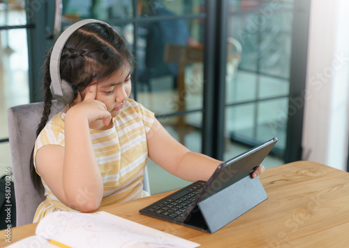 Little girl in headphones thinking while doing her homework on digital tablet.Kid learning class study online video call zoom with laptop.Home schooling,Online learning concept.