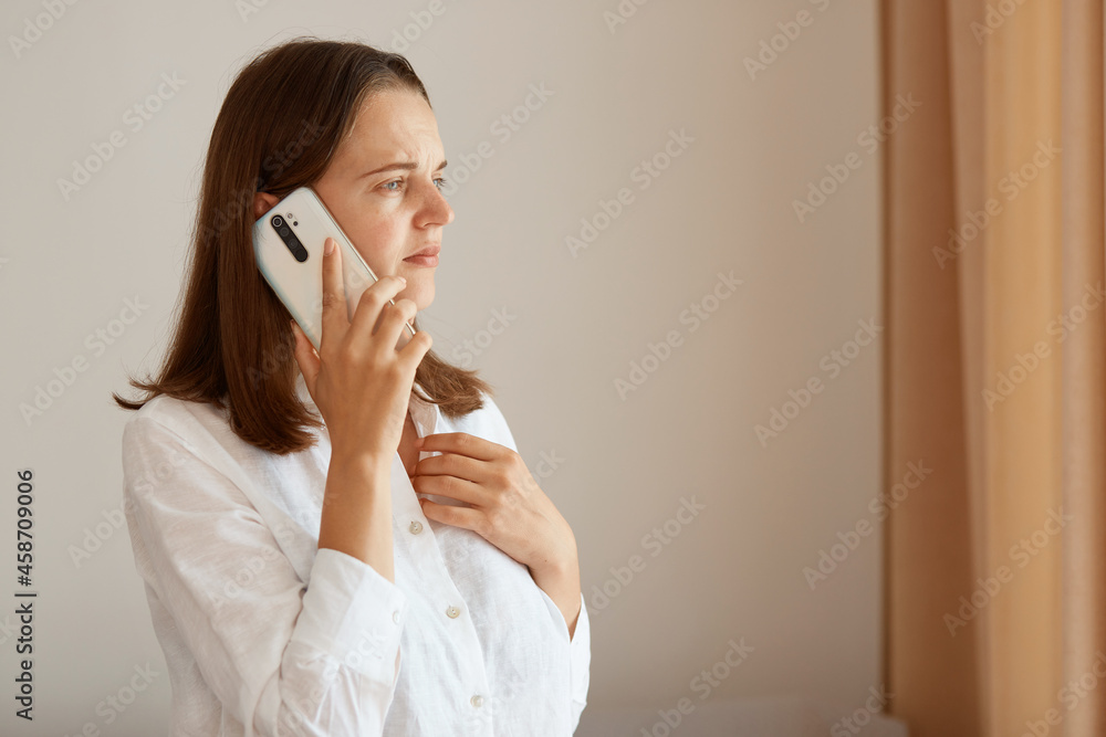 Portrait of upset female with dark hair wearing white cotton shirt standing indoor and talking phone, hearing bed news, having sad facial expression, looking away. Portrait of upset female with dark hair wearing white cotton shirt standing indoor and talking phone, hearing bed news, having sad facial expression, looking away.