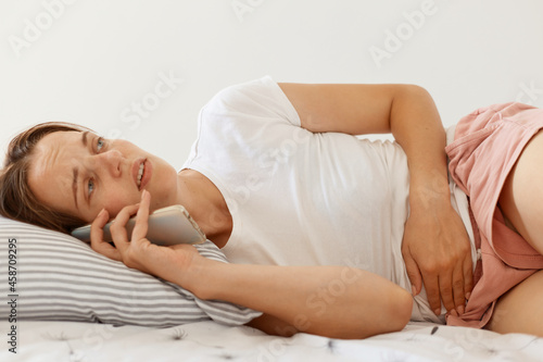 Ill woman with dark hair wearing white casual t shirt lying in bed and talking via smart phone with her doctor, having problems with stomach, suffering from abdominal pain.