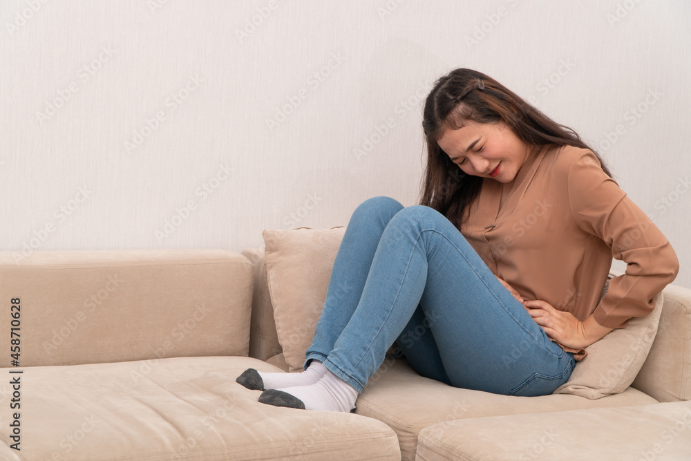 Unhappy Asian woman sitting on the sofa and holding on stomach suffering. Abdominal pain that comes from menstruation, diarrhea, or indigestion. Sickness and healthcare concept Unhappy Asian woman sitting on the sofa and holding on stomach suffering. Abdominal pain that comes from menstruation, diarrhea, or indigestion. Sickness and healthcare concept