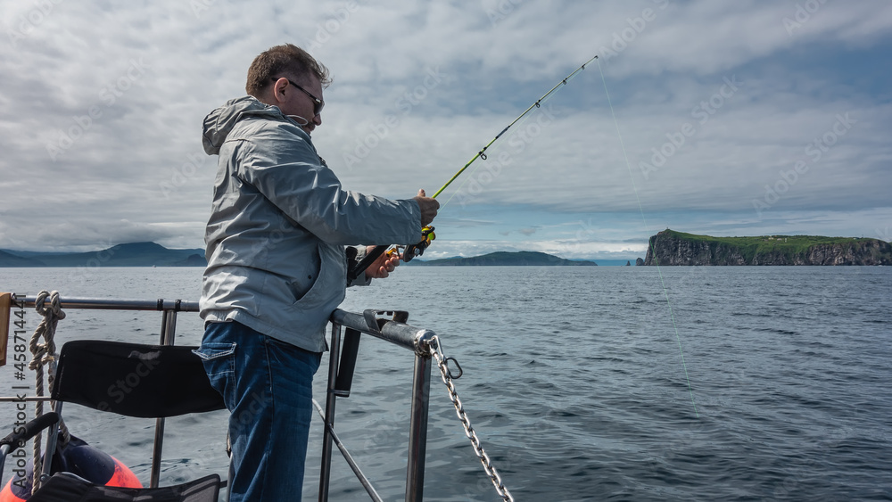 A male fisherman stands on board a yacht with a fishing rod in his hands. The fishing line is submerged in water. Picturesque rocks against a background of blue sky and clouds. Kamchatka A male fisherman stands on board a yacht with a fishing rod in his hands. The fishing line is submerged in water. Picturesque rocks against a background of blue sky and clouds. Kamchatka