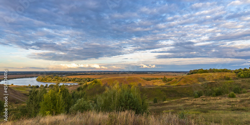 The vibrant colors of the sunset over the hill. Colorful landscape in the evening, gorgeous blue sky with golden clouds over a field with hills and a river. A magical moment of nature. © Sergei