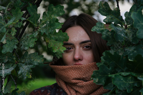 beautiful young woman with red hair looks out from behind the leaves of a tree in the park in autumn. High quality photo