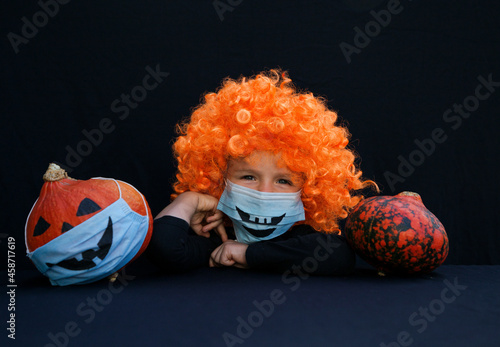 Fototapeta Naklejka Na Ścianę i Meble -  boy 4 years old in orange wig and pumpkin in blue medical mask with a painted scary smile. On black background. Halloween decorations and celebrations in new reality of the COVID-19 pandemic.