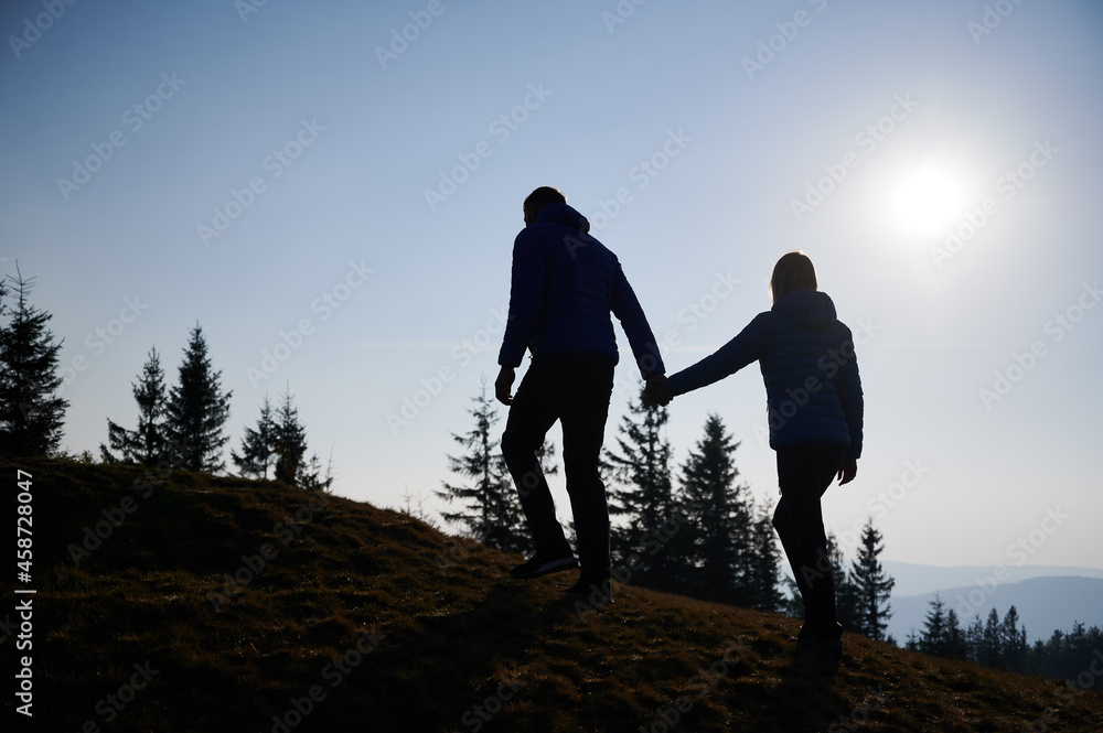 Back view of young man holding his wife's hand and leading her while they climbing on mountain hill during walk in fresh air. Trekking trip in the autumn mountains.