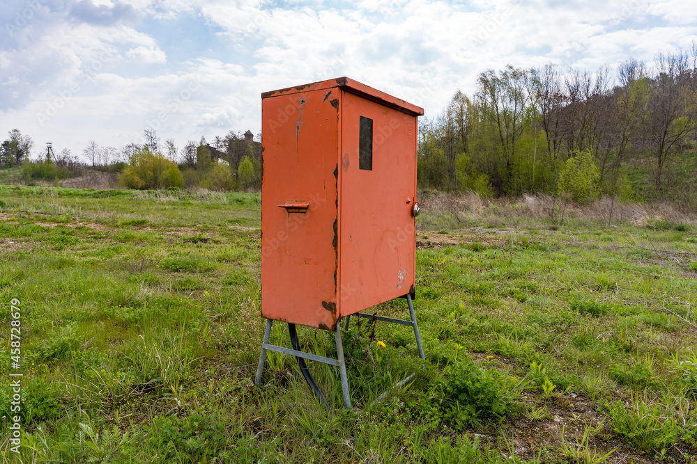 Orangener Elektrokasten oder Verteilerkasten aus Metall auf einer grünen Wiese vor einer Baustelle. Orangener Elektrokasten oder Verteilerkasten aus Metall auf einer grünen Wiese vor einer Baustelle.