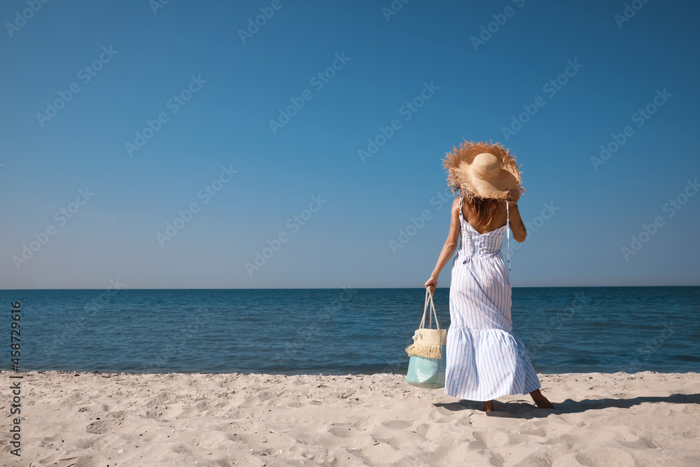 Woman with beach bag and straw hat on sand near sea, back view Woman with beach bag and straw hat on sand near sea, back view