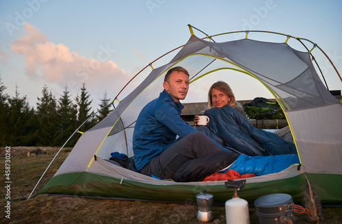 Smiling woman who just woken up with her husband in tent after sweet night in the open air, holding metal cup in her hand. Hikers watching amazing view of the morning mountains.