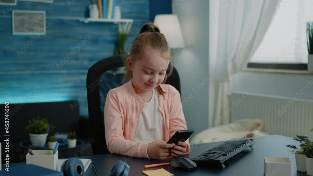 Young girl using smartphone with touch screen at desk. Little child smiling while looking at phone screen after class lesson and school education. Schoolgirl having fun on modern device Young girl using smartphone with touch screen at desk. Little child smiling while looking at phone screen after class lesson and school education. Schoolgirl having fun on modern device