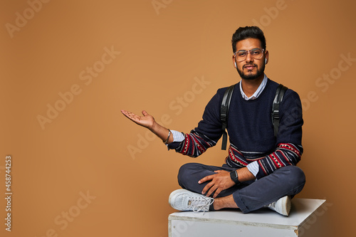 Young indian man sitting on background and pointing with backpack