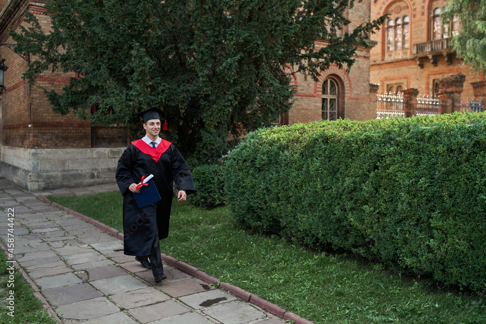 Happy male graduate walking in university campus in graduation robe, copy space for text