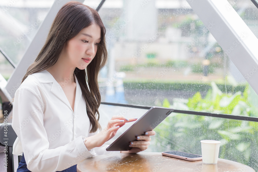 Young Asian business woman is looking at tablet or notepad in her hands while she is sitting at coffee shop among business background in the city. Young Asian business woman is looking at tablet or notepad in her hands while she is sitting at coffee shop among business background in the city.