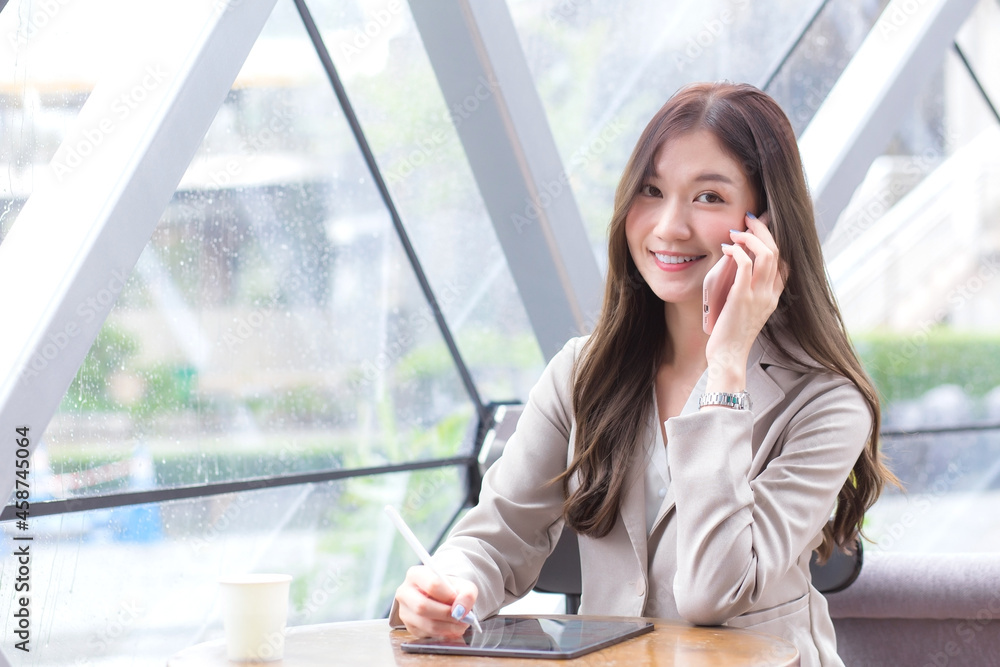 Asian professional businesswoman in a cream-colored suit holds a smartphone to chat and record information on a tablet while in a coffee shop. Asian professional businesswoman in a cream-colored suit holds a smartphone to chat and record information on a tablet while in a coffee shop.