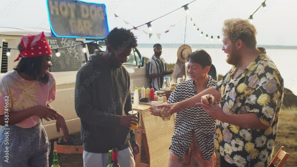 Group of cheerful multiethnic friends chatting, laughing and clinking glasses in toast while standing by food truck at outdoor summer festival Group of cheerful multiethnic friends chatting, laughing and clinking glasses in toast while standing by food truck at outdoor summer festival