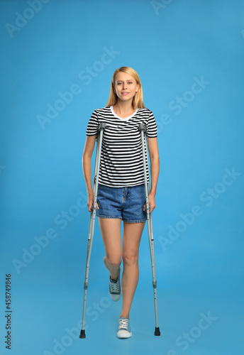 Young woman with axillary crutches on light blue background