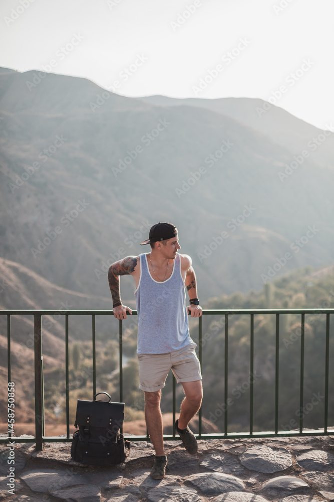 Young caucasian guy with a tattoo all over his arm stands near railing at the observation deck in the mountains. Scenic view in Armenia. High quality