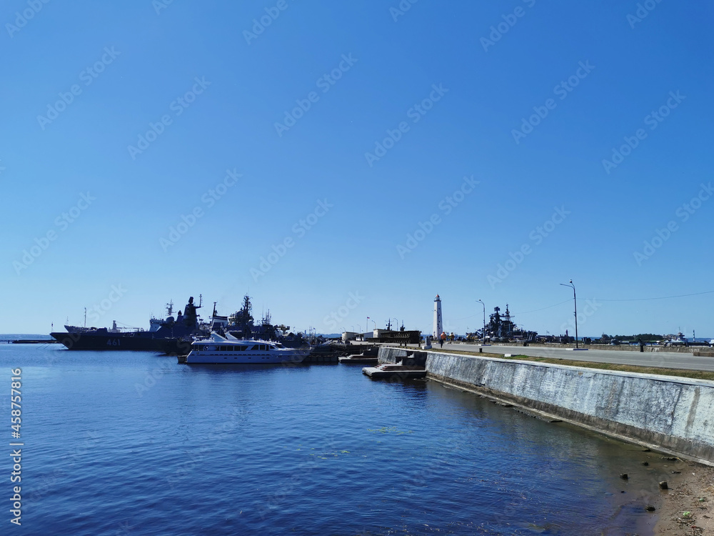 View of warships, boats and a pier with a lighthouse from Petrovskaya Embankment against the blue sky on a summer day in Kronstadt.