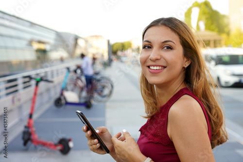 Close-up of confident young business woman turns around and smile using smartphone app she is preparing to use electric scooter to go back home