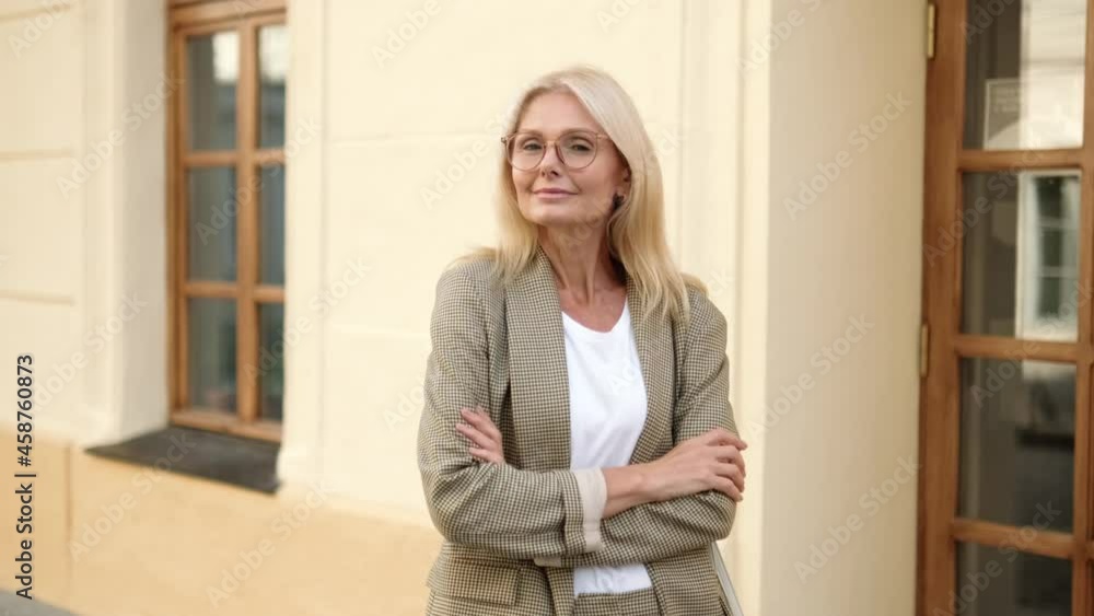 Smiling blonde mature businesswoman posing to the camera standing near building on the street Smiling blonde mature businesswoman posing to the camera standing near building on the street