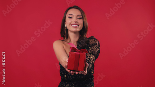 happy and blurred woman smiling and holding gift box on red.
