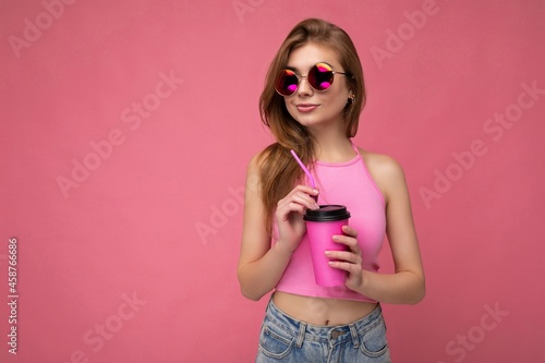 Charming young happy blonde woman wearing stylish clothes isolated over colourful background wall holding paper cup for mockup drinking milkshake looking to the side