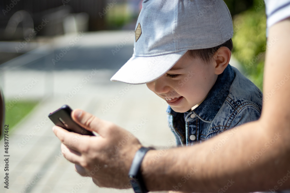 Dad is showing the phone to the smiling kid with rare teeth. Happy family, young dad and cute little boy child son are spending time outside. Blurry background. Dad is showing the phone to the smiling kid with rare teeth. Happy family, young dad and cute little boy child son are spending time outside. Blurry background.