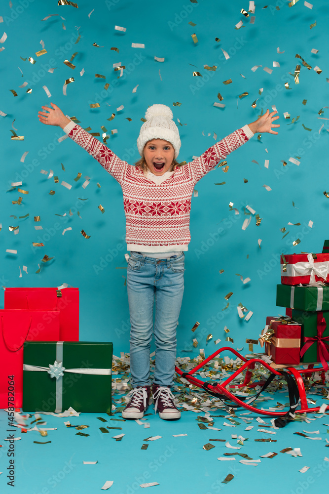 Obraz premium stylish and astonished kid with outstretched hands near sled and gift boxes on blue