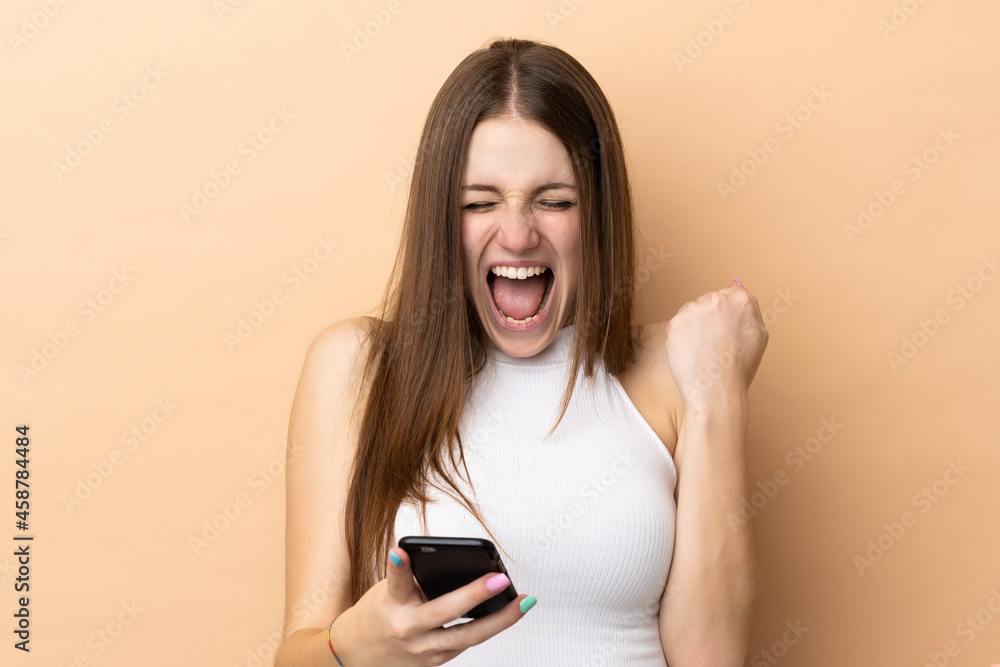 Young caucasian woman isolated on beige background with phone in victory position Young caucasian woman isolated on beige background with phone in victory position