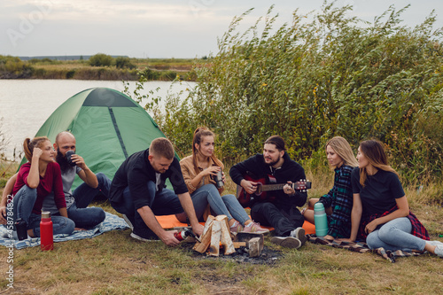 A group of friends sit by the fire on the background of the river and sing to the guitar. © kanashkin