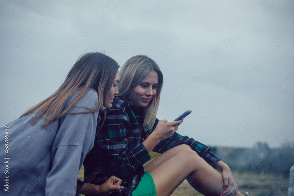 Obraz premium Couple of girls sitting in nature cute chatting using smartphone.
