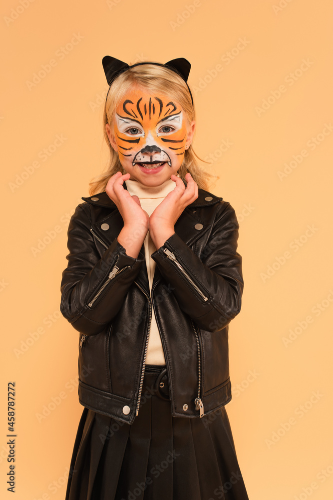 smiling kid in black leather jacket and tiger makeup holding hands near face isolated on beige smiling kid in black leather jacket and tiger makeup holding hands near face isolated on beige