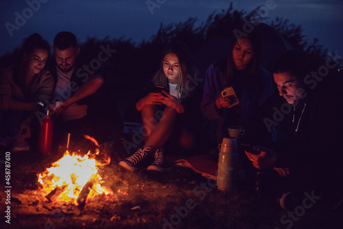 A group of friends are sitting around a campfire at night using smartphones.