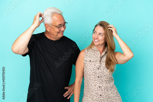 Middle age couple isolated on blue background having doubts while scratching head
