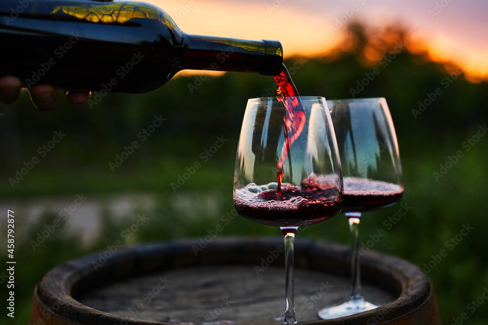 Pouring red wine into glasses on the barrel at dusk - obrazy, fototapety, plakaty 