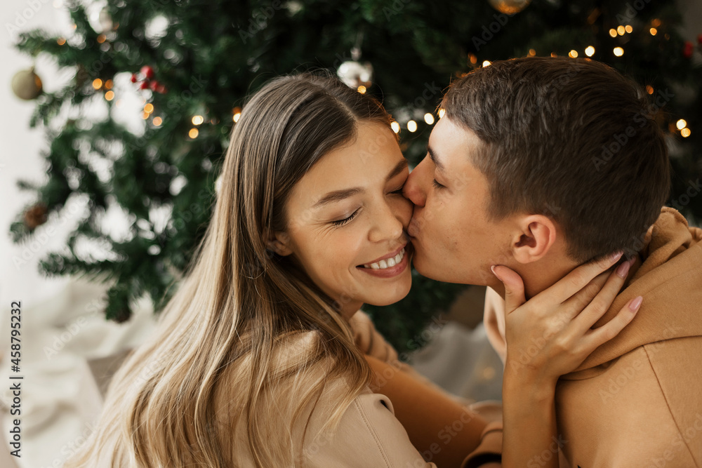 Young couple sitting near Christmas tree at home. Man kissing a woman. Love and relationships. Christmas and New Year celebration Young couple sitting near Christmas tree at home. Man kissing a woman. Love and relationships. Christmas and New Year celebration