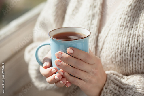 A woman in a warm cardigan holds a blue mug in her hands. Close-up of female hands holding cup of coffee or tea.