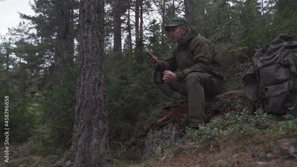 The traveler sits on a halt in a coniferous forest, looks at the phone and drinks from a metal mug The traveler sits on a halt in a coniferous forest, looks at the phone and drinks from a metal mug