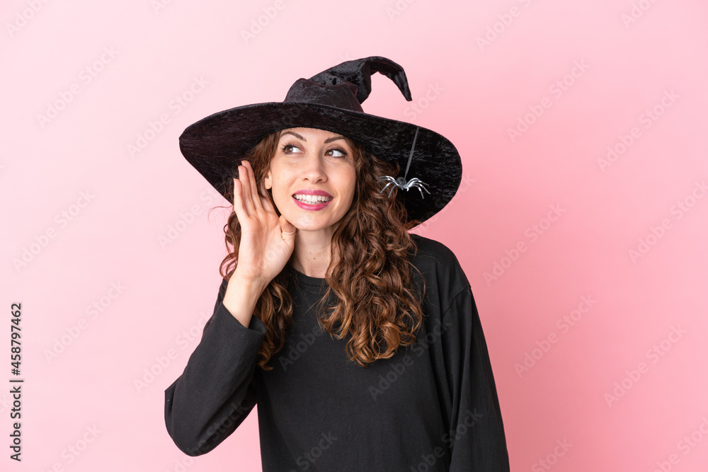 Young caucasian woman celebrating halloween isolated on pink background listening to something by putting hand on the ear