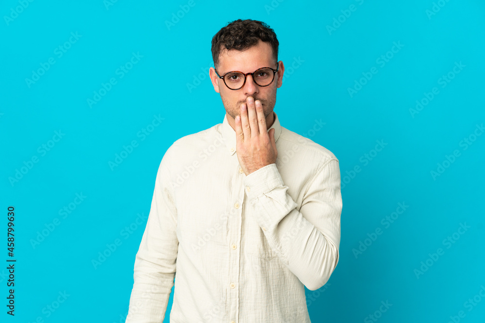 Young caucasian handsome man isolated on blue background covering mouth with hand