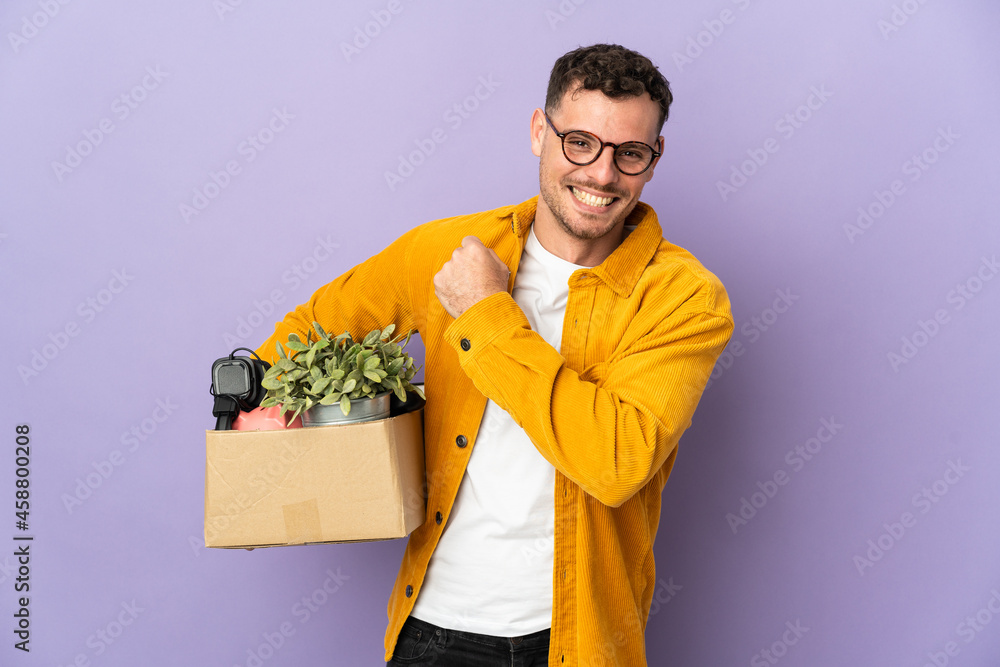 Young caucasian man making a move while picking up a box full of things isolated on purple background celebrating a victory Young caucasian man making a move while picking up a box full of things isolated on purple background celebrating a victory