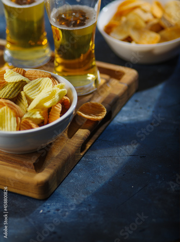 Amber beer in glasses and a bowl of potato chips on a wooden tray. Low angle view. Blue background. Rest, relaxation in the company of friends, watching TV and sports matches.
