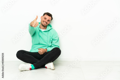 Young handsome caucasian man sitting on the floor with thumbs up because something good has happened