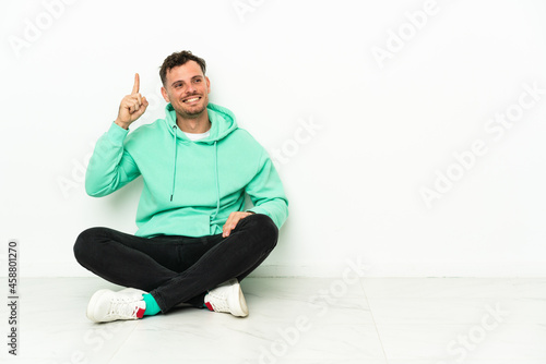 Young handsome caucasian man sitting on the floor intending to realizes the solution while lifting a finger up