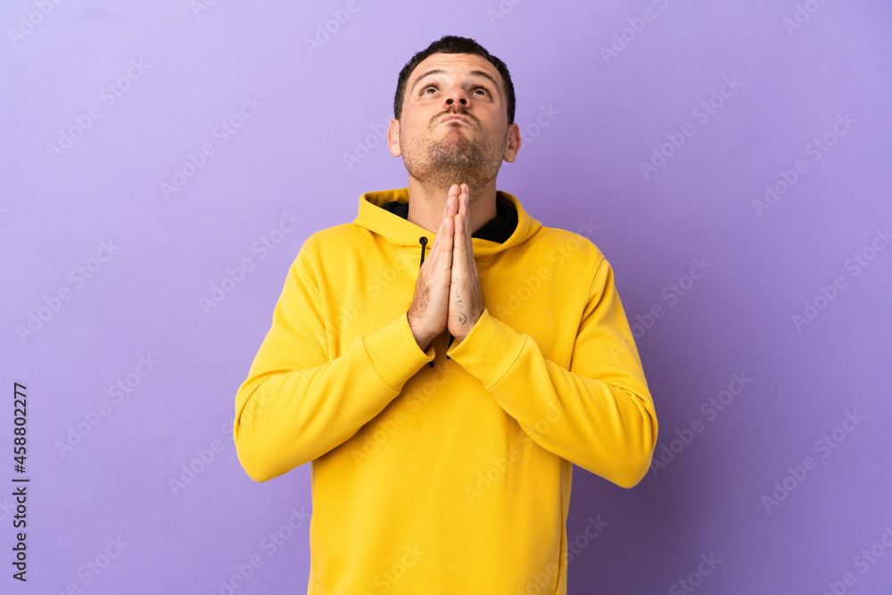 Brazilian man over isolated purple background keeps palm together. Person asks for something Brazilian man over isolated purple background keeps palm together. Person asks for something