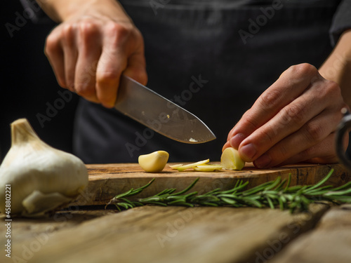The chef cuts the garlic on a cutting board. A sprig of rosemary. Wooden texture. Macro photography. Hot spice for salads, sauces, soups, meat, fillings. Restaurant, hotel, cafe, home cooking.