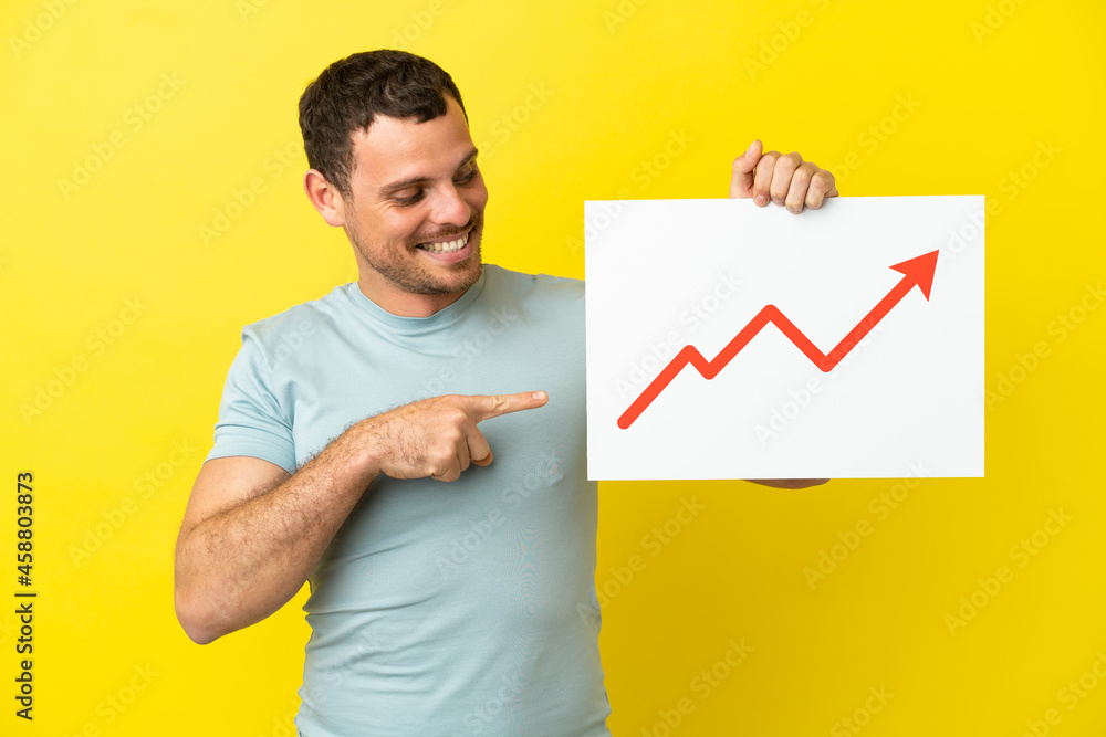 Brazilian man over isolated purple background holding a sign with a growing statistics arrow symbol and pointing it Brazilian man over isolated purple background holding a sign with a growing statistics arrow symbol and pointing it
