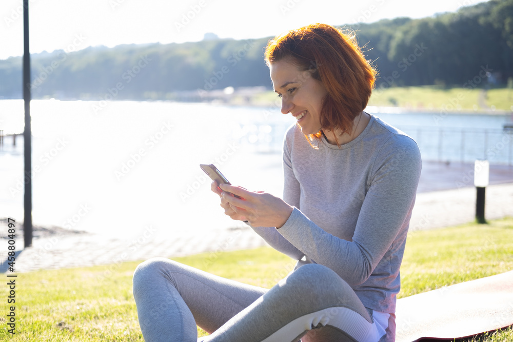 Naklejka premium slim ginger woman in sport clothes using mobile phone at park. Health, wellness, fitness lifestyle