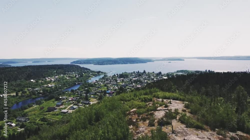 smooth flight over a mountain on a drone, on which there are large green trees, in the distance a small town with small houses, a river flows into the lake, small islands on the lake, the sky is blue, smooth flight over a mountain on a drone, on which there are large green trees, in the distance a small town with small houses, a river flows into the lake, small islands on the lake, the sky is blue,